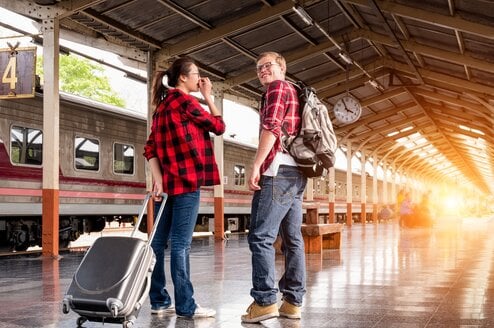 a man with a backpack and a woman with a suitcase, both wearing red flannel shirts, prepare to leave for their train